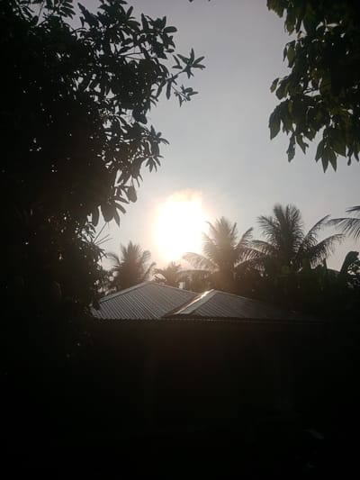 A sunburst is visible in the sky, seen through the leaves of trees. The sun is high in the sky, with its rays fanning out. In the foreground, the corrugated metal roof of a building is visible, with a dark structure below it. Palm trees are seen in the background, silhouetted against the bright sky. The image appears to be taken in a tropical or subtropical location. It is not possible to determine the specific location within Kruengbaroblangme, Indonesia from the image alone. There are no people or animals visible in the image.