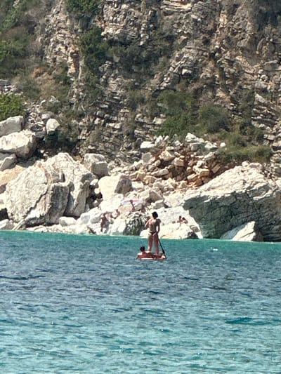 The image depicts a rocky coastline with clear turquoise water. Two people are visible in the water. One person, wearing a dark swimsuit, is on a stand-up paddleboard, while another person is in the water nearby, possibly swimming. The sun appears to be shining, creating sparkling reflections on the water's surface. The rocky shore is covered with sparse vegetation, suggesting a warm, possibly Mediterranean climate. The scene is likely taking place in the daytime, given the bright lighting. The overall impression is one of a leisurely outdoor activity in a scenic coastal environment. There is no visible text in the image.
