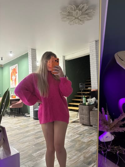 A person takes a selfie in a mirror. The person is wearing a bright pink, sweater dress. The mirror reflects the surrounding room, which includes a staircase, artwork, furniture, and decorative items. The location may be in the city of Большая Шильна, Russia.