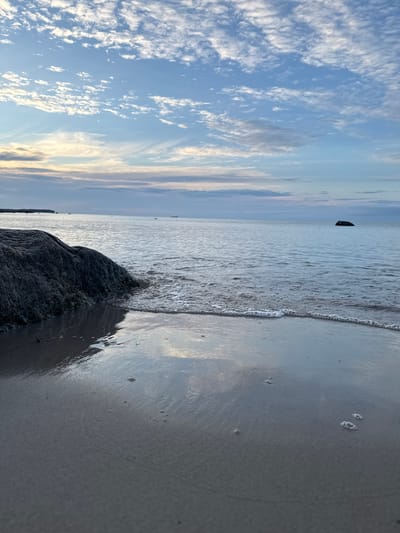 This is a vertical, eye-level, medium shot of a beach at sunset. The scene is calm and serene, with soft light illuminating the sky and water.
The subjects of the image are the natural elements of a beach: the sky, the water, and the sand. The sky is a pale blue, dotted with wispy, white clouds that are tinged with pink and gold from the setting sun. The water is a tranquil, shimmering expanse, with small waves gently lapping at the shore. The sand is wet and reflective, with small pockets of air creating a delicate bubbly texture near the water's edge. A large, dark rock is prominently positioned in the foreground, anchoring the composition. In the distance, a dark, indistinct object appears to be floating on the water, possibly a buoy or a distant boat.
The time of day is sunset, indicated by the warm hues in the sky and the soft, diffused light. The weather appears to be clear and calm, with no strong winds or visible precipitation. The location is a beach, likely in Estonia given the context. The overall mood of the image is peaceful and contemplative, inviting the viewer to relax and appreciate the beauty of nature. There is no visible text in the image.