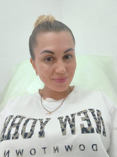 A Caucasian woman with blonde hair pulled into a bun, wearing a white sweatshirt with the words "HOY NEW" and "NWOTIMWOO" printed on it, is looking at the camera. She is wearing a necklace with a charm. Her left ear is visible with a small hoop earring. The background is a plain white wall and a light green sheet. The image appears to have been taken indoors. There is no information in the image to suggest a specific location in Komsomolsk-on-Amur, Russia.