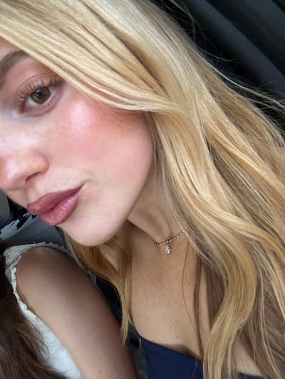 The image is a close-up, tilted portrait of a person, likely female, with blonde, wavy hair. Her head is angled, revealing her right eye, part of her nose, and her lips, which are slightly parted with a hint of a smile and appear to have pink lip gloss. Her right eye is brown, and she has visible freckles on her fair skin, particularly on her cheek and neck area.

She is wearing a delicate gold-colored necklace with a small, teardrop-shaped clear stone or charm around her neck, and a dark blue garment is visible. In the bottom right corner, a portion of another person or object with brown hair and a white, possibly lacy, garment can be seen. The background on the top left features a dark, textured surface, suggestive of a car interior. The lighting is bright and even. No text is visible. The location is Saint-Victor-la-Coste, France.