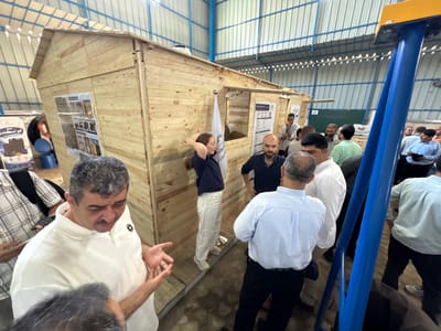 In the city of Gaza, Palestinian Territories, a group of men and one woman are gathered around a small, unfinished wooden structure resembling a house, located inside what appears to be a warehouse or workshop. The structure is made of light-colored wooden planks and has a pitched roof. There are posters or informational displays attached to the exterior walls of the structure.

In the foreground, a man in a white polo shirt with a textured pattern is gesturing with his hands as he speaks to someone out of frame. To his left, another man wearing a plaid shirt is visible. Behind the wooden structure, a woman in a dark blue shirt and white pants is standing with her back to the camera, her hand touching her hair. Several other men are also present, observing the structure or engaged in conversation. Some are wearing casual shirts and trousers, while others are in more formal attire.

The environment suggests an indoor industrial or exhibition space, with metal roofing and ribbed metal walls visible in the background. A blue metal structure, possibly part of an exhibit or piece of equipment, is prominent on the right side of the image. The lighting appears to be from overhead sources, illuminating the scene.