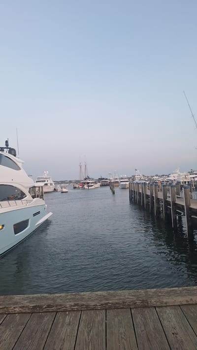 The image depicts a serene marina scene in Nantucket, United States of America, likely during twilight. The foreground is dominated by a weathered wooden dock, its planks showing signs of age and use, with a dark, rippling body of water stretching out before it. Several boats are moored in the water. On the left, a large, modern light blue and white yacht is prominently featured, its sleek design evident. Further back, a variety of other vessels are visible, including smaller motorboats and at least one classic sailing ship with tall masts. The boats are docked along wooden piers that extend into the water. In the background, a hazy shoreline with buildings can be seen, suggesting a populated area. The sky above is a pale, clear blue, transitioning to a lighter hue near the horizon, indicating the setting or rising of the sun. No people are clearly visible in the image, and the overall atmosphere is one of calm and quiet.