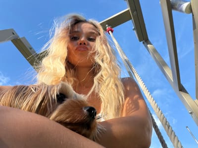 An overhead, low-angle, wide shot captures a young blonde woman holding a small, tan dog against a bright blue sky. The woman is positioned in the lower center of the frame, looking directly at the camera with her lips pursed. Her long, wavy blonde hair is windswept. The dog, which appears to be a Yorkshire Terrier, is held in her arms, close to her face. The background consists of a clear blue sky with some wispy clouds and the metallic structure of what appears to be a playground or swing set, with thick ropes hanging down. The image was taken outdoors on a sunny day in Thep Krasatti, Thailand.