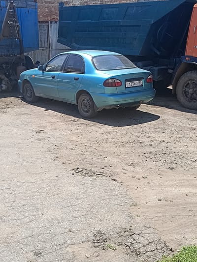 In Berdiansk, Ukraine, a teal blue sedan is parked on a rough, unpaved surface, possibly a yard or an industrial area, with visible cracks and debris. Behind the sedan, a large blue dump truck, with its bed raised, is prominently positioned, and a portion of an orange truck is visible to the right. The backdrop includes a brick wall and corrugated metal fencing. The car's license plate reads "C133AO 185 RUS". The lighting suggests it is daytime.