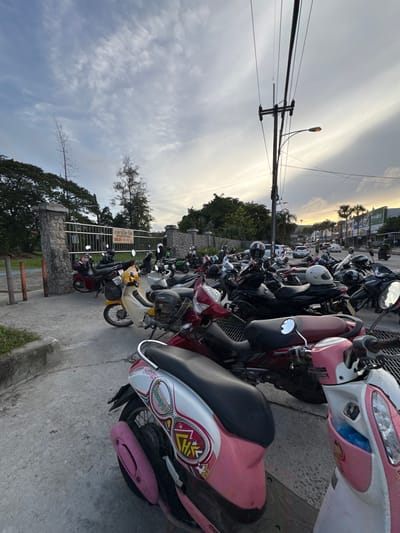 This media file depicts a row of parked motorcycles and scooters on a concrete surface, likely a parking lot or street. The scene is set outdoors, with a backdrop of trees and some buildings visible in the distance. The sky is overcast with streaks of light suggesting either sunrise or sunset. The time of day appears to be late afternoon or early evening.

The subjects are primarily the vehicles, which are predominantly scooters, with a few motorcycles interspersed. Many of the scooters are brightly colored, with prominent examples being pink and white, and yellow. Several of the scooters have helmets resting on them or attached. There are no people visible in the frame, but the presence of the vehicles suggests they are nearby.

Notable details include a street lamp visible in the distance, and a metal fence with a stone base to the left. A sign with Thai text is affixed to the fence. The overall atmosphere is one of a typical urban or semi-urban scene in Southeast Asia, likely Thailand, given the location context of Phuket City Municipality. The weather appears to be mild, with no strong indicators of rain or extreme heat. The lighting suggests a diffused light, common during twilight hours. The vehicles are parked in a somewhat orderly fashion, indicating a designated parking area.