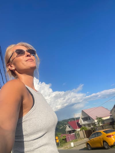 In this outdoor medium shot, a blonde woman with sunglasses and a pearl earring is captured from a low angle, looking upwards towards a vibrant blue sky dotted with fluffy white clouds. She is wearing a grey tank top. To her right, a street lined with houses and a yellow car is visible. The scene appears to be in a hilly area, with lush green trees and more houses visible in the distance. The sunlight suggests it is a bright, clear day. The image is oriented vertically. No visible text is present in the image.