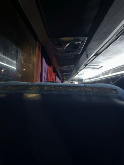The image is a low-angle, interior shot taken from the back of a bus, looking forward. The foreground is dominated by the top of a dark blue seat. Above the seat, the bus interior is visible, with the ceiling and the sides of the bus. On the left side, a window shows a reflection and outside lights. Along the right side of the ceiling, there are various fixtures, including vents and control panels, and in the distance, more seating and people can be seen. The scene appears to be during nighttime, with artificial lighting present inside the bus. The overall environment suggests a travel setting within a bus.