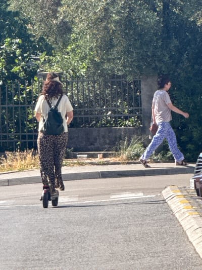 In Donji Pobori, Montenegro, a person on an electric scooter with curly brown hair and a white t-shirt, wearing leopard print pants and a dark backpack, is moving away from the camera on a paved road. To the right, a second person wearing a light-colored t-shirt and blue patterned pants is walking on a sidewalk. The background consists of green foliage and a black metal fence. The scene is illuminated by sunlight.