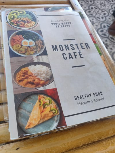 The image is a close-up, overhead shot of a menu on a bamboo table. The menu features several food items, including a smoothie bowl with fruit and granola, a dish with smoked salmon, boiled eggs, and greens, a bowl of rice with curry, and a folded crepe filled with fruit. The menu also displays text that reads "Live, Love, Eat. DON'T WORRY, BE HAPPY", "MONSTER CAFÉ", and "HEALTHY FOOD Meanam Samui". The background shows a patterned floor. The setting is likely a cafe or restaurant in Baan Tai, Thailand, as indicated by the text on the menu. There are no people or animals visible in the image.