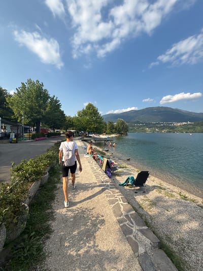 A woman with dark hair pulled back walks on a gravel path towards the right of the frame, wearing a white t-shirt, black shorts, and a light gray backpack. She is seen from behind and her legs are visible. The path is lined with planters containing green foliage on the left and a low stone wall on the right, separating it from a pebbled beach. Several people are visible near the edge of a large, calm body of water. Some are seated on the shore, while others are in the water. The water is clear and reflects the bright blue sky, which is dotted with white, fluffy clouds. In the background, a tree-covered mountain range with a small town visible on its slope rises above the lake. The scene appears to be a lakeside campground or recreational area, likely in Calceranica al Lago, Italy, given the mountainous backdrop and lake setting. Two black lounge chairs are set up on the pebbled beach, one with a green towel draped over it.