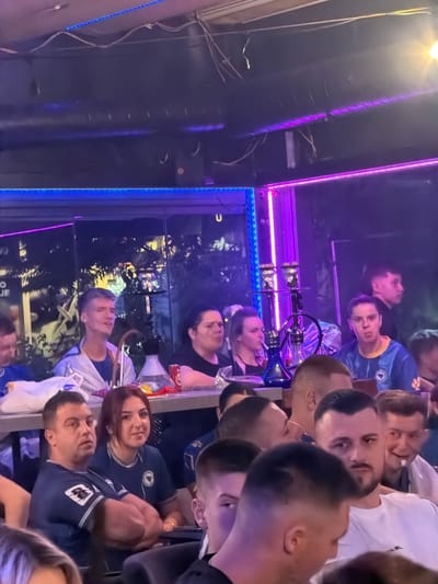 The image depicts an indoor gathering of people, likely in a bar or lounge. The environment is dimly lit, with vibrant neon lighting, predominantly blue and purple, illuminating the space. Several individuals are seated at tables, some engaged in conversation, while others appear to be observing something. A prominent hookah is visible, suggesting a hookah lounge setting. The people in the image are diverse in age and gender. Some individuals are wearing sports-themed attire. The overall atmosphere appears relaxed and social. There is no discernible visual information indicating the specific location in the city of Travnik, Bosnia and Herzegovina.