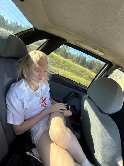 A young woman with blonde hair is sitting in the back seat of a car. She is wearing a white t-shirt with a red logo and white shorts. She has her eyes closed and a slight smile on her face, with sunlight casting shadows on her. The car's interior is visible, including grey fabric seats and the interior of the rear window. Outside the window, a green, hilly landscape with trees under a clear blue sky can be seen. The image appears to have been taken in a rural or suburban area outside the city of Sosnovyi Bor, Russia, given the landscape.
