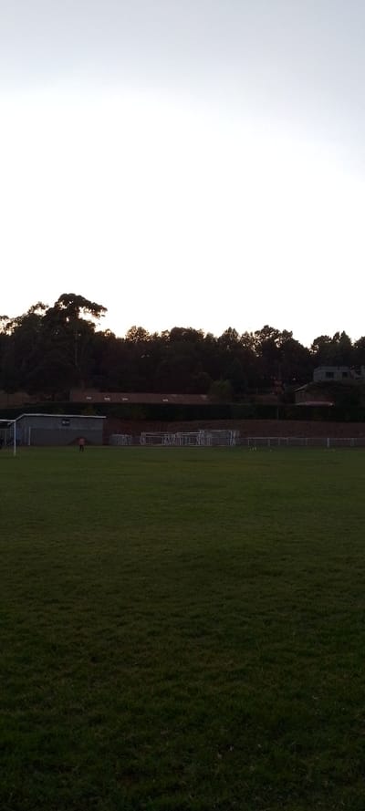 A wide, verdant field dominates the foreground. A solitary figure stands near a gray building, possibly a shed, beside a soccer goal. Rows of equipment line the perimeter. A raised brick wall separates the field from a line of dark trees. Above the trees, a few structures, including a building with a gable roof, are visible. The sky is a pale, overcast white, suggesting twilight or dusk.