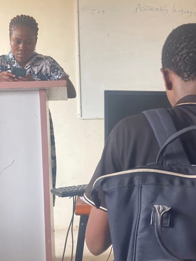 The scene depicts an interior setting, likely a classroom or computer lab, within the city of Okigwe, Nigeria. In the foreground, viewed from behind, is a person with dark skin and short, dark hair, wearing a black short-sleeved shirt with white trim on the collar and sleeves. A dark blue backpack with light grey straps is worn over their shoulders. This individual is seated at a wooden desk, facing a dark computer monitor, with a black keyboard positioned on the desk. Visible to the left, slightly further back, is a woman with dark skin and dark braided hair, wearing a patterned short-sleeved blouse. She is leaning against a light-colored lectern or stand with a reddish-pink trim along its top edge. Her gaze is directed downwards at a dark-colored mobile phone she holds with both hands. In the background, a white whiteboard is mounted on a light-colored wall, displaying handwritten text in blue marker: "CDE" and "Assembly language". Cables are visible beneath the desk where the foreground subject is seated. No direct interaction between the two individuals is evident.