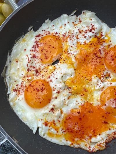 A close-up, overhead shot captures a frying pan filled with six fried eggs, generously sprinkled with red pepper flakes. The eggs are sunny-side up, with their bright orange yolks still liquid and glistening. The whites are firm but slightly browned at the edges. The pan is dark and non-stick, with a visible rim. In the upper left corner, a corner of a clear plastic container holding some yellow, oval-shaped items, possibly potatoes or pickles, is visible. The lighting is bright, casting a slight glare on the yolks. The overall impression is a vibrant and appetizing breakfast dish, prepared in a home kitchen, most likely in Beşiktaş, Turkey, given the common breakfast preparations in that region. There are no people or any other objects clearly identifiable in the scene, nor any specific environmental context beyond what is implied by the presence of a frying pan and food.