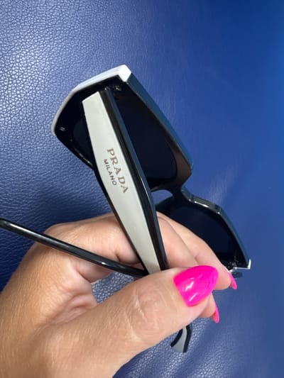 A hand with bright pink fingernails is holding a pair of black and white Prada sunglasses against a textured blue background. The sunglasses are angled, with the Prada Milano logo visible on the white arm. The image is a close-up shot, focusing on the sunglasses and the hand. There is no indication of the location being Orsk, Russia.