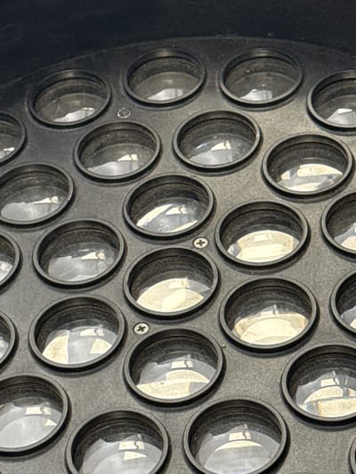 The media file displays a close-up, detailed view of a light fixture with numerous circular lenses arranged in a grid pattern. The fixture appears to be made of dark, possibly metal material, with a textured surface. Each lens is recessed within its own circular housing, and they reflect light, creating bright highlights and darker shadows within their surfaces. Some of the reflections suggest an indoor environment with artificial lighting.
There are no people or discernible activities taking place. The focus is entirely on the object itself and its intricate design.
Notable details include the consistent grid arrangement of the lenses, the presence of small screws or fasteners visible between some of the lenses, and the reflective quality of the glass surfaces. The lighting suggests it could be daytime or nighttime, as the light source is internal to the fixture and reflecting off its components. No specific location cues from Baku City, Azerbaijan, are visible in the image itself. No text is present in the image.