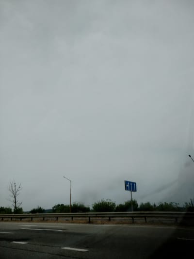 The image shows a road with guardrails and trees. A road sign indicating multiple lanes and directions is visible on the right side of the road. The sky is overcast, and the overall lighting suggests a cloudy or foggy day. The scene appears to be along a highway, potentially in a rural or suburban area, given the presence of trees and the open road. The location is identified as Krasiv, Ukraine.