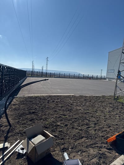 The image displays an outdoor industrial or commercial site in Pliska, Bulgaria, under a clear, bright blue sky. In the foreground, a section of dark, disturbed earth is visible, prepared for landscaping or construction. Located on this soil are a cardboard box containing light-colored rectangular blocks, several silver metal gate rack segments, and a silver spray can. A black corrugated conduit extends from the left edge of the frame, running along the base of a fence and curving upwards into a large, dark gray paved area. A concrete curb separates the soil from the paved surface.
In the midground, the expansive, empty paved area stretches towards the horizon. Along its left perimeter, a dark metal fence with a decorative, geometric pattern runs continuously. In the far midground and background, multiple tall utility poles and lattice towers support a series of high-voltage power lines that traverse the sky. On the far right, a portion of a light-colored industrial building with multiple windows is visible, adjacent to a large metal lattice tower. The distant horizon reveals a faint, hazy outline of a low mountain range or hills. No individuals are present, but the visible materials indicate ongoing or recently completed construction or installation work.
