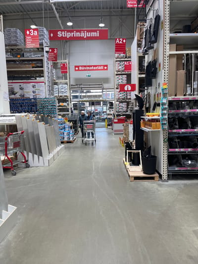 The image captures a wide aisle within a hardware or home improvement store, located in Liepaja, Latvia. The store features extensive shelving units on both sides, stocked with a diverse array of products. The floor is a light grey concrete, and the lighting suggests an indoor, daytime setting.
In the mid-ground, at least one person is visible pushing a shopping cart down the aisle, engaging in typical retail shopping activity. Another red shopping cart, possibly containing large format tiles or similar building materials, is positioned in the foreground on the right.
The shelves display a wide variety of goods. On the left, large bags, likely containing building mixes or insulation, are stacked on pallets. Further down this aisle, smaller items and various hardware components are arranged. On the right, black pipe segments and other heating or ventilation components are prominent, alongside displays of what appear to be floor tiles.
Red signs with white text, written in Latvian, are prominently displayed throughout the aisle, indicating product categories and section numbers. Visible text includes:
* Large overhead signs: "Stiprinājumi" (Fasteners) and "Būvmateriāli" (Building materials).
* Aisle/section indicators: "A2" and "A1".
* Product categories: "Kamīnkrāsnis" (Fireplaces/Stoves), "Apkures" (Heating), and "Eļļas radiatori" (Oil radiators).
* Detailed lists under signs "3a" and "3": "Naglas, Skrūves, Dībeļi, Paplāksnes, Uzgriežņi" (Nails, Screws, Dowels, Washers, Nuts) and "Hermētiķi, Hidroizolācija, Būvkalumi, Auklas, ķēdes, Troses" (Sealants, Waterproofing, Building hardware, Ropes, chains, Cables).
A "DEPO" logo, identifying the store chain, is also visible on equipment in the background.