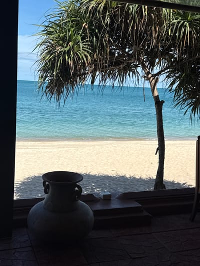 The image depicts a serene beach scene in Baan Lamai, Thailand, viewed from an interior space. In the foreground, a large, dark, round ceramic vase with two handles sits on a textured, dark floor. Behind the vase, a wooden ledge runs horizontally, casting a shadow from a tree. The tree, with its distinctive spiky fronds, is a prominent feature, partially obscuring the view of the beach and the ocean. The beach itself is covered in light-colored sand, with gentle waves lapping at the shore. The ocean is a vibrant turquoise, stretching out to the horizon under a clear blue sky with scattered white clouds. The overall impression is one of tranquility and natural beauty, enhanced by the framing of the scene through the interior space. There are no people or animals visible in the image.