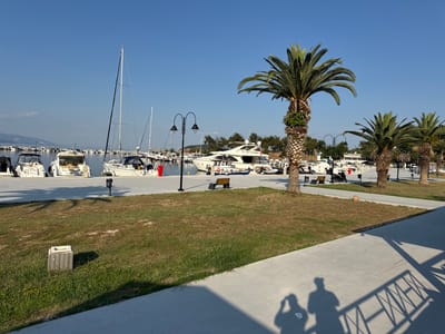 The image depicts a marina in Nea Irakleitsa, Greece, under a clear blue sky. Several yachts and sailboats are moored in the water. A paved walkway runs along the waterfront, bordered by a grassy area with palm trees. In the foreground, a shadow of two people can be seen on the walkway, cast by the bright sunlight. A concrete planter and a bench are visible in the grassy area.