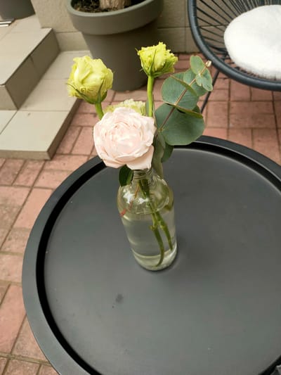 A close-up, overhead shot shows a clear glass bottle containing a bouquet of flowers and eucalyptus leaves. The bottle rests on a round, dark gray table. The bouquet consists of one light pink rose in full bloom and two pale yellow roses that appear to be budding. Several large, oval, silvery-green eucalyptus leaves are also present. The background reveals a reddish-brown tiled patio, a light gray concrete planter, and a portion of a dark-colored chair with a white cushion. The scene appears to be an outdoor setting. The image was taken in Kremenchuk, Ukraine.
