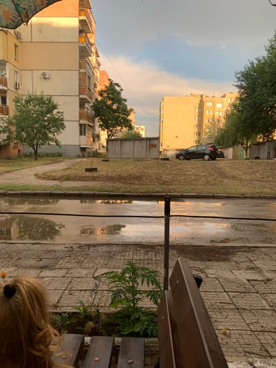 The image depicts an outdoor scene in Vidin, Bulgaria, likely a residential courtyard, during what appears to be late afternoon or early evening. The foreground shows the back of a person with blonde hair, seated on a wooden bench, facing away from the camera. Beside the bench are some small plants, one with purple flowers, and a small orange object, possibly a flower. The ground in the immediate foreground is paved with square concrete tiles, some of which are wet and reflective, suggesting recent rain.
Beyond the paved area, a large puddle of water is visible, reflecting the sky and surrounding buildings. A dark, thin horizontal line, possibly a hose or cable, runs across the puddle. Further back, a grassy area with sparse vegetation and a few trees is present. The middle ground features a concrete wall and a black car parked on the grass.
In the background, two apartment buildings are visible. The building on the left is taller and appears to be in a state of disrepair, with peeling paint and exposed concrete. The building on the right is lighter in color and shows several windows. The sky is partly cloudy, with the setting sun casting a warm glow on the buildings. The overall impression is one of a somewhat rundown but lived-in urban environment.