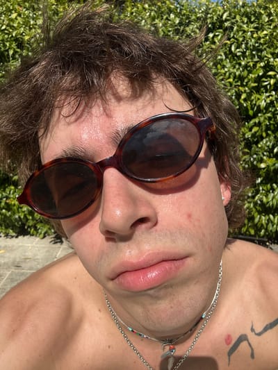 A close-up, tilted, portrait shot shows a young man lying on his back, possibly outdoors, evidenced by greenery in the background. The man is fair-skinned and wearing round, dark-lensed sunglasses with brown tortoise-shell frames. He has brown hair, an earring in his left ear, and a silver necklace with beads. On his upper right chest, there's a tattoo of abstract design in black. The man appears to be lying near a stone or concrete surface, possibly a patio or walkway. The lighting is bright, suggesting it is daytime. The image suggests relaxation in an outdoor setting. There is no immediate visual evidence to definitively place the image in Lourmarin, France, beyond the potential for its resemblance to landscapes from the general area.