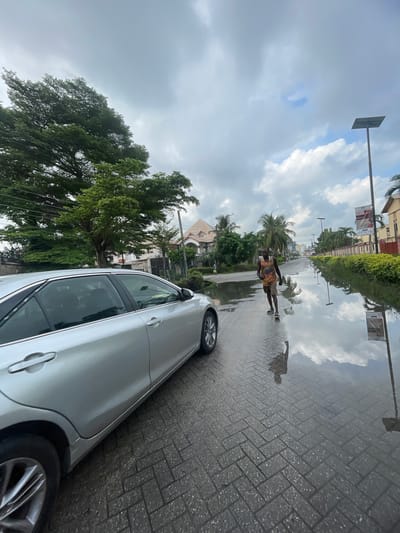 A street scene in Moba, Nigeria, following rain, features a silver car parked partially on a wet, patterned-brick sidewalk. A man is walking through a puddle on the sidewalk. Reflections of the sky, buildings, and greenery are visible in the standing water on the ground. Overhead, overcast skies with a few breaks in the clouds are visible. Streetlights and foliage are seen lining the street.
