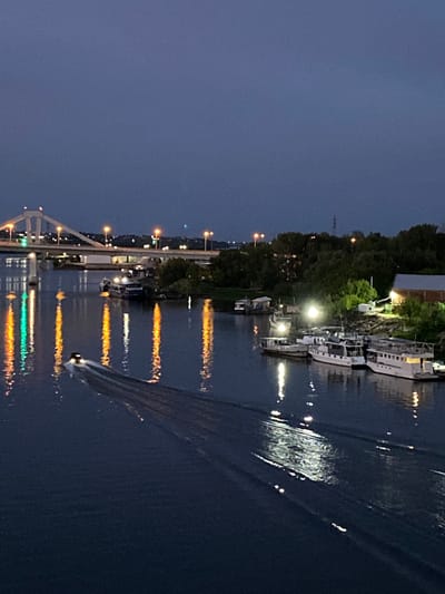 This image depicts a dusky twilight scene over a body of water, likely the Kondurcha River, near Novokuybyshevsk, Russia. A prominent arched bridge, illuminated by scattered lights, spans the water in the background. Several boats are moored along the riverbank on the right, with lights reflecting on the dark water. One motorboat is actively moving across the water in the foreground, leaving a wake. The shoreline is lined with trees, and a building with illuminated windows is visible on the right bank. The sky is a deep, darkening blue, indicating the transition from day to night. The overall atmosphere is calm and serene.