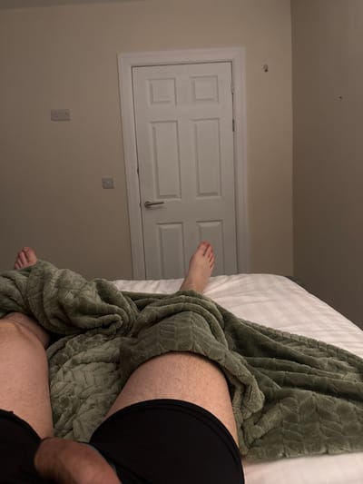 The scene depicts an interior room, consistent with a bedroom. A subject is positioned supine on a bed, covered by white, vertically striped linen and a textured, sage-green blanket. The perspective is first-person, looking towards the subject's feet and the room beyond. The subject's bare feet are visible above the blanket. The upper thighs and groin area are visible, displaying body hair. Black boxer briefs are worn, with a penis exposed and resting above the garment. In the background, a white, four-panel door with a brushed metal handle is centered. To the left of the door, a double electrical outlet is mounted on the light-colored wall, with a single switch or outlet below it. The wall to the right of the door exhibits minor surface imperfections. The specific geographical location within Southampton, United Kingdom, is not discernable from the visual information provided.