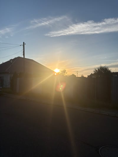 A wide-angle, low-angle, over-the-shoulder shot depicts a sunset over a residential area, with the sun's rays flaring across the lens. In the foreground, a dark asphalt road curves to the left, with a manhole cover visible in the lower right corner. A curb separates the road from a sidewalk. Beyond the sidewalk, a low, light-colored fence runs horizontally across the frame, separating the street from the properties. A house with a brown tiled roof is partially visible on the left, with a tall utility pole with multiple wires in front of it. In the background, trees and the roofs of other houses are silhouetted against the bright sky. The sky is a gradient of pale blue at the top, transitioning to a hazy yellow-orange near the horizon, with thin, wispy white clouds. The overall impression is one of a peaceful evening in a suburban setting. The specific location is the vicinity of Osiedle Słoneczne in Szczecin, Poland, as indicated by the characteristic architecture and streetscape of this district.