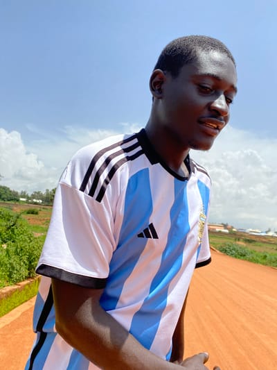 The image shows a young Black man, possibly of Nigerian descent, wearing a blue and white striped soccer jersey with black trim and the Adidas logo. He is looking up and to his left with a slight smile, his eyes closed. The jersey appears to be for the Argentine national soccer team, suggested by the blue and white stripes and the emblem on the chest.
The man is outdoors, with a bright blue sky and scattered white clouds behind him. To his right, there is a glimpse of a reddish-brown dirt surface, possibly a road or a field, and some green vegetation and buildings in the background. The lighting suggests it is daytime, likely late morning or early afternoon, given the brightness of the sun and the shadows.
The man's expression suggests a moment of enjoyment, perhaps during a sports activity or simply relaxing outdoors. The image is taken from a low angle, emphasizing the sky and the man's face. There is no visible text in the image.