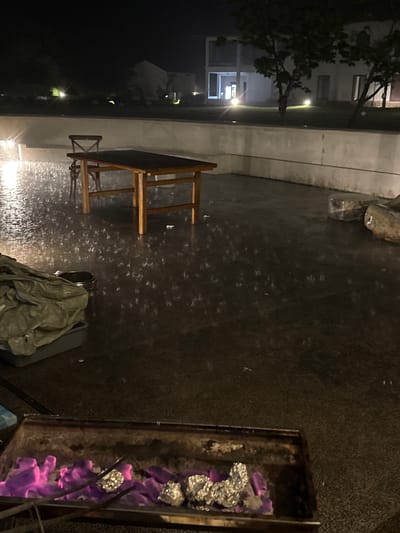 Night view of a patio in Jinhua, China, showing a wooden table and chairs next to a low, white wall. Rain falls on the dark patio surface. A grill with glowing purple coals covered in foil sits in the foreground. A covered garbage can is near the table. In the background, a house is lit by streetlights.