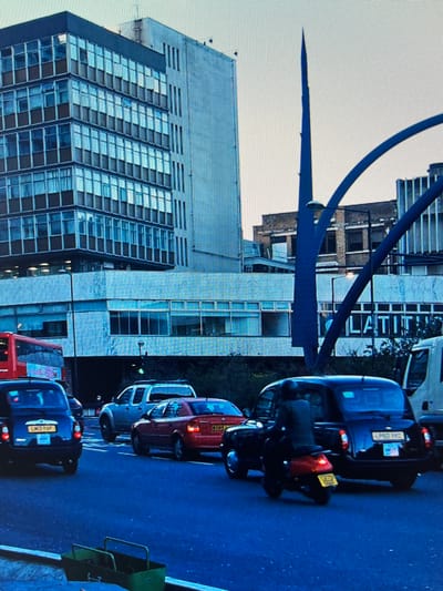 The image depicts a street scene with several vehicles and buildings. In the foreground, a red bus is visible, and behind it, a blue van with a license plate reading "LT11 VCP." Further down the street, a dark-colored car with illuminated brake lights and a license plate is parked. A red car is also visible, parked next to a white van.
The background features a modern, multi-story building with a glass facade, reflecting the sky and surrounding structures. Adjacent to this is an older, brick building with visible windows. A large, dark blue, curved structure arches over the scene, possibly a bridge or an architectural element. The overall impression is an urban environment, potentially during daylight hours due to the bright sky, though the lighting on the street suggests it might be overcast or late afternoon. The image appears to be taken from a slightly elevated perspective, looking down the street. There are no visible people in the frame. The scene is likely a city street, possibly in Europe given the style of the vehicles and architecture.