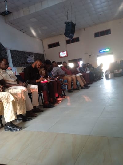 A group of seated people, mostly men, are attentively listening or participating in an event. They are dressed in a variety of attire. Some are taking notes, while others have their hands clasped. The venue seems to be a hall or church, evident from the speaker system hanging from the ceiling. There is natural light streaming in from a doorway or window. The floor is tiled.