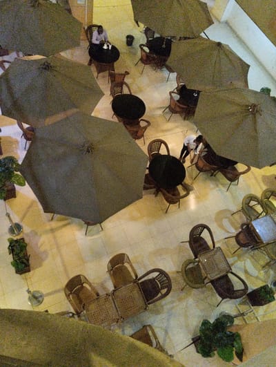 An aerial view reveals an outdoor cafe or restaurant setting, partially covered by large khaki-colored umbrellas. Underneath, tables and chairs, some occupied, are visible. A person in a white shirt is seen near one table. Tile flooring dominates the visible ground. Potted plants are scattered around, adding touches of greenery. The scene has a slightly muted, perhaps nighttime, ambience.