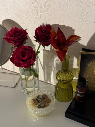 The image shows a still life composition with flowers and decorative objects. In the foreground, three deep red roses are arranged in a clear glass vase, with their stems and some green leaves visible. To the left of the roses, a single dried flower with reddish-brown petals and yellow accents is positioned, casting a shadow on the textured white surface behind it.
Behind the roses and dried flower, two olive-green glass bottles with fluted designs are placed horizontally. One bottle has a narrower neck and appears to be filled with a clear liquid. The other bottle is wider.
Further back, on a dark tray with a gold embossed pattern, a small brown bottle with a black cap is visible. Text on the bottle reads "GROWN ALCHEMIST BODY CREAM CREME POUR LE CORPS".
To the right of the bottles, a small, ornate clear glass dish contains several smooth, multi-colored stones. The overall lighting suggests it might be artificial light, possibly from a lamp, creating soft shadows and highlighting the textures of the objects and the background surface. The scene is indoors, likely on a shelf or table, given the arrangement of items. No people are visible, and the mood is calm and decorative. The location context is provided as Rossiyskiy, Russia, but there are no specific visual cues in the image to confirm this.