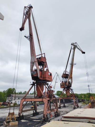 This image depicts an active industrial port or loading facility in Medvezhyegorsk, Russia, under an overcast sky during daytime. The scene is dominated by several large, heavy-duty gantry cranes, notably aged and covered in rust, indicating their extensive use.

In the foreground and center, two prominent cranes are engaged in bulk cargo handling. The crane on the left features a large, open clamshell grab bucket suspended over extensive piles of dark, granular material, likely coal or ore, which lie adjacent to multiple railway tracks. This crane's robust base rests on its own set of tracks. The second large crane, located towards the right, is captured mid-action, emptying its bucket into the open hold of a ship or barge, a segment of which is visible on the right side of the frame. A smaller, more distant gantry crane is also visible between these two main structures.

The ground area around the cranes and tracks is a mix of dirt, gravel, and spilled bulk material. In the background, beyond the industrial operations, a grey, multi-windowed industrial building stands amidst green trees and foliage, with a smaller yellow industrial vehicle (possibly a front loader) visible in the distance.

Notable identification marks include the number "52" visible on the left crane (both on its upper structure and lower chassis) and the number "43" on the second large crane, likely serving as unit identifiers. The overall atmosphere is functional and utilitarian, reflecting the heavy industrial nature of the port operations. No people are visible in the image.