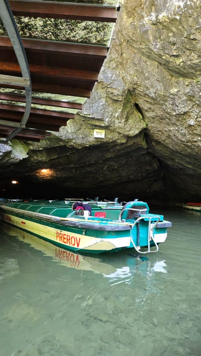 This media file shows an interior view of a cave with a boat tour. The scene is set within a rocky cave, with a wooden staircase descending to the water's edge on the left. The cave walls are rough and uneven, with some patches of green moss. The staircase has metal railings and wooden steps.
A green and yellow boat with a white stripe is docked in the clear, green water. The boat has a motor at the back and what appears to be seating for passengers. The word "Přerov" is visible in red lettering on the side of the boat, with its reflection also visible in the water. There is a small yellow sign on the cave wall with some text, possibly indicating a name or warning. Two lights are visible further into the cave, illuminating the darkness. A person's torso is visible leaning over the boat, possibly a tour guide or passenger. The overall impression is of an underground boat tour. The lighting suggests it could be daytime, but the enclosed nature of the cave makes it difficult to determine the exact time of day or weather. The location is identified as Vavřinec, Czechia.
