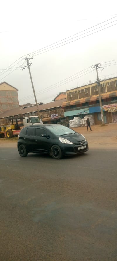 A black Honda Fit is parked on the street in front of buildings on a cloudy day. The car has black rims and the license plate is visible. Across the street is a man walking on the sidewalk. A yellow bulldozer and a white delivery truck are parked on the side of the road. Overhead are several telephone poles with wires extending to the buildings.