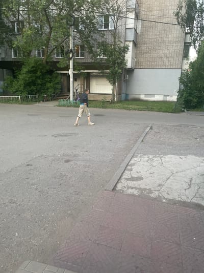 A person walks across an asphalted courtyard in front of a multi-story residential building in Shebunino, Russia. The building has light-colored brickwork and several windows, with trees and foliage surrounding its base. The person, wearing a dark jacket and light-colored shorts, is walking towards the right side of the frame. The ground is a mixture of asphalt and concrete, with some patchy areas and visible manhole covers. A green metal fence and a ramp leading to the building's entrance are also visible in the background. The lighting suggests it is daytime, possibly late afternoon or early evening, with overcast conditions.