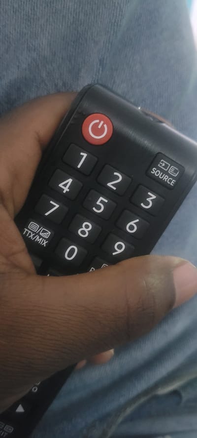 The media file shows a close-up of a person's hand holding a black remote control. The hand is dark-skinned and appears to be resting on a light blue, textured surface, likely denim. The remote control has a red circular button with a white power symbol on it, located on the left side. Above the power button are several numbered keys, ranging from 1 to 9 and 0. At the top of the remote, there is a rectangular button labeled "SOURCE" with a small icon above it. Towards the bottom of the remote, there is a button labeled "TTX/MIX" with a similar icon. The person's thumb is partially visible, resting on the remote control. The scene is lit, and the background is out of focus, suggesting a casual indoor setting. The image provides a detailed view of the remote control and the hand holding it, with no clear indication of the time of day, weather, or specific emotions. The location context of Kipala Mpakani, Tanzania, is provided externally.