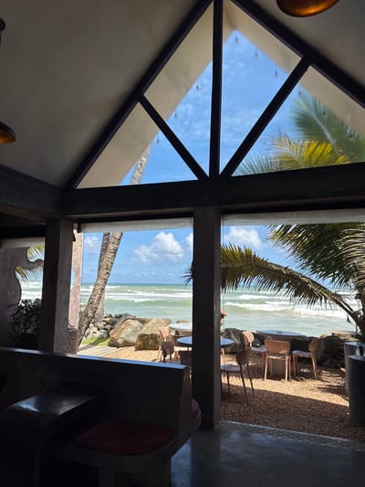 This is a daytime, eye-level, interior shot of a restaurant or cafe in Ahangama, Sri Lanka, with a view of the ocean through large open archways and a triangular skylight. The foreground is dominated by dark, built-in seating made of concrete or stone with red cushions. Through the opening, palm trees are visible, along with large rocks and the ocean with waves breaking on a rocky shore. In the mid-ground, outdoor seating is arranged on a gravel-covered area. Several light-colored chairs are placed around round tables, some with white tablecloths. The sky is blue with scattered white clouds. The architecture features exposed concrete beams and a pitched roof with triangular glass sections. The overall impression is a tropical, open-air dining experience.