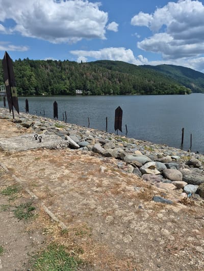 The image depicts a rocky shoreline on a body of water, likely a lake or reservoir. In the foreground, there is a patch of dry, sandy ground with sparse vegetation and a concrete slab with two metal rings. Beyond this, a dense collection of various sized rocks forms the bank of the water. Several dark, pointed metal posts are embedded in the rocky shore and in the water, some appearing to be remnants of a pier or mooring structure. The water itself is a deep blue-gray, reflecting the cloudy sky. Across the water, a heavily wooded hillside rises, dotted with trees in various shades of green, with some patches of dry or discolored foliage visible. A small, white building with a dark roof is situated on the hillside. The sky above is partly cloudy with large, white cumulus clouds against a bright blue background. The scene appears to be located in Pancharevo, Bulgaria, likely a recreational area along a lake. There are no people visible in the image.