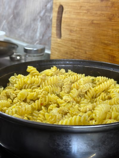 A close-up, eye-level shot shows a generous portion of rotini pasta mixed with what appears to be scrambled eggs or finely crumbled chicken, coated in a creamy, yellow sauce and seasoned with herbs. The dish is served in a large, dark-colored pot with a slightly reflective surface. In the background, out of focus, are kitchen elements including a stainless steel canister with a silver lid, a dark grey metal bowl, and a portion of a wooden cutting board with a vertical, dark handle. The lighting is dim, creating shadows and highlighting the texture of the food. The scene is set in a kitchen environment, likely in the city of Crokhovo, Russia, given the context provided. No people are visible in the image.