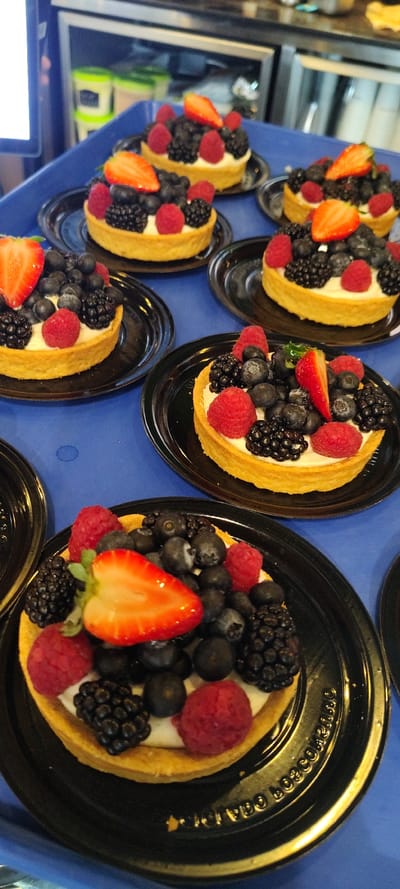 The image displays an arrangement of multiple individual fruit tarts, neatly placed on a blue surface, likely a tablecloth or tray. Each tart consists of a golden, fluted pastry shell filled with a creamy white base, generously topped with a colorful assortment of fresh berries. The berries include bright red sliced strawberries, plump red raspberries, dark blue blueberries, and glossy black blackberries.

The tarts are served on small, oval-shaped black plastic plates. Along the rim of these plates, embossed text is visible, which appears to read "PREMIUM QUALITY Appetizer Cup" or similar phrasing, with words like "QUARTER SIZE" also partially discernable.

In the background to the left, there are blurred reflections on what appears to be a glass or metallic surface, possibly from a refrigerator or display case, along with a partial view of a laptop screen in the lower-left corner. The scene suggests a prepared display of desserts, ready for serving or consumption at an event or gathering, possibly in Cuernavaca, Mexico, under indoor lighting conditions.